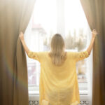 Young,Woman,Opening,Window,Curtains,At,Home,,Back,View